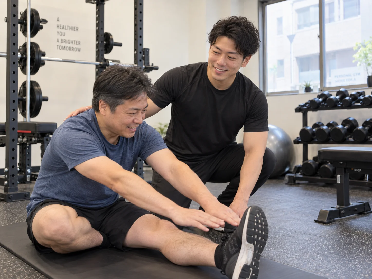 座って脚を伸ばすストレッチを、トレーナーがそばでサポートする様子
